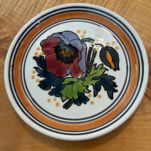 May include: A round ceramic plate featuring a hand-painted floral design in the center with a large purple and red poppy flower, green leaves, and a closed bud. The rim is decorated with a wide orange band and thin black circular lines.