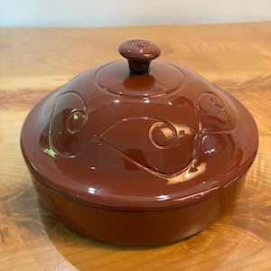 May include: A round, glossy, dark brown ceramic lidded dish. The lid features a raised, embossed swirling pattern and a small, circular knob handle at the center. The dish has a smooth, reflective finish and a simple, cylindrical base design.