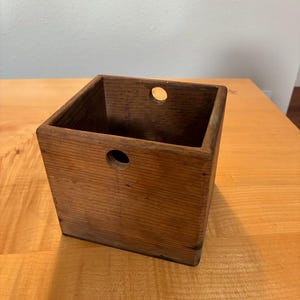May include: A square, open-top wooden box featuring a medium brown wood grain finish. The front and back panels each contain a single, centered circular hole near the top edge, providing a clear view through the interior of the container.