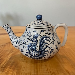 May include: A white ceramic teapot featuring a hand-painted blue rooster motif surrounded by intricate scrolling vine and leaf patterns. The teapot includes a matching lid with a blue knob, a curved handle, and a tapered spout, all decorated with blue.