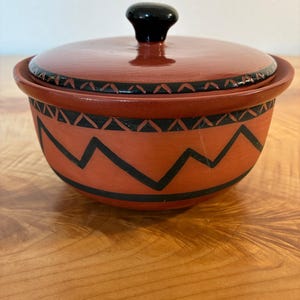 May include: A terracotta-colored ceramic bowl with a matching lid featuring a black knob. The bowl and lid are decorated with hand-painted black geometric patterns, including a zigzag line around the center and a repeating leaf-like motif.