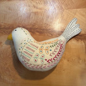 May include: A cream-colored felt bird ornament featuring a yellow beak and intricate hand-stitched embroidery in shades of red, pink, orange, green, and blue. The decorative patterns include floral motifs, chevron lines, and dashed details.