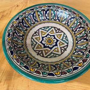 Może przedstawiać: Okrągła ceramiczna miska z ręcznie malowanym geometrycznym wzorem gwiazdy pośrodku i motywem kwiatowym. Projekt łączy żywe odcienie turkusu, głębokiego błękitu, żółci i fioletu na kremowym tle z grubym turkusowym obrzeżem.
