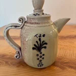 May include: A cream-coloured ceramic teapot with a crackled glaze finish, featuring a dark blue hand-painted floral motif and the word "Tae" on the front. It has a curved handle with a scroll detail, a matching lid, and a short, angled spout.