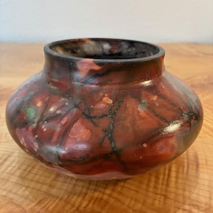 May include: A small, round ceramic vase with a wide opening and a smooth, glossy finish. The surface features a mottled pattern of deep red, burnt orange, and black tones, accented by thin, dark, web-like crackle lines throughout the exterior.