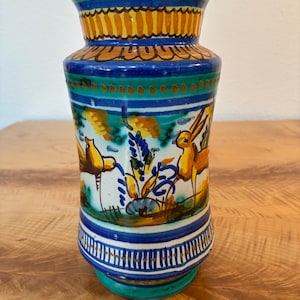 May include: A ceramic vase featuring a hand-painted design with yellow deer, blue floral motifs, and green accents. The neck and base are decorated with horizontal bands of yellow, blue, and white stripes, creating a colorful, patterned appearance.