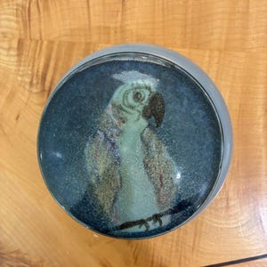 May include: A circular decorative object featuring a glossy, dark blue surface with a speckled texture. Centered on the surface is a stylized illustration of a light-colored parrot with a dark beak, perched on a thin, dark horizontal line.