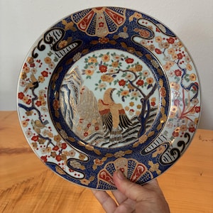 May include: A round ceramic plate featuring a central illustration of a bird perched near rocks and flowering branches. The design includes a deep blue border with gold accents, fan-shaped motifs, and floral patterns in red, white, and gold colors.