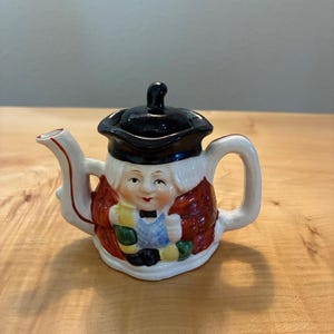 Könnte beinhalten: Eine Keramik-Teekanne in Form einer Figur mit schwarzem Dreispitz, rotem Mantel und blauer Weste. Die Teekanne hat einen weißen Ausguss, einen weißen Henkel und ein bemaltes Gesicht mit schwarzer Fliege. Der schwarze Deckel bildet den Hut.