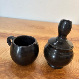May include: A black ceramic cup with a rounded body and a small handle, positioned next to a black ceramic lidded vessel featuring a bulbous base and a narrow, elongated neck. Both items have a smooth, dark finish and a simple, functional design.