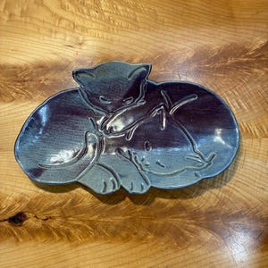 May include: A ceramic dish in an oval shape featuring a relief design of two curled-up cats. The piece has a glossy glaze with a color gradient transitioning from deep purple to a muted blue-grey, highlighting the feline forms and their tails.