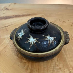 May include: A dark brown ceramic lidded pot with two side handles and a central knob on the lid. The lid features three white, star-shaped leaf patterns painted on its surface. The pot has a smooth, glazed finish with a lighter tan rim.