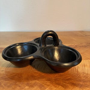 May include: A black ceramic serving dish featuring three interconnected circular bowls arranged in a triangular formation. A single arched handle is positioned in the center, connecting the three bowls, which have smooth, rounded rims and deep interiors.