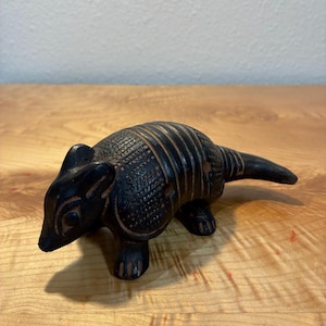 May include: A dark brown, carved wooden armadillo figurine featuring detailed etched patterns on its shell, pointed ears, and a long tail. The surface of the wood shows natural grain variations and deep, textured grooves across the body.