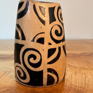 May include: A cylindrical vase with a light tan base colour featuring a bold, black hand-painted pattern of interconnected spirals and geometric semi-circular shapes that wrap around the exterior surface of the vessel.