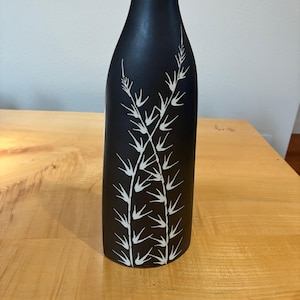 May include: A matte black ceramic vase with a narrow neck and a cylindrical body. The front surface features a hand-painted white botanical design consisting of two vertical stalks with pointed, leaf-like branches extending outward.