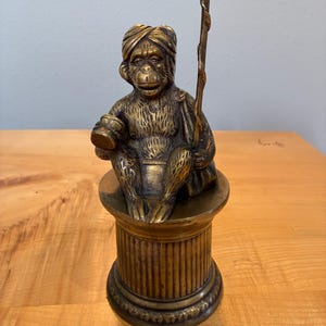 Könnte beinhalten: Eine Messingfigur eines sitzenden Affen mit Turban, der eine kleine Trommel in der einen und einen langen Stab in der anderen Hand hält. Der Affe sitzt auf einem zylindrischen, kannelierten Sockel mit einem dekorativen Rand am Fuß.