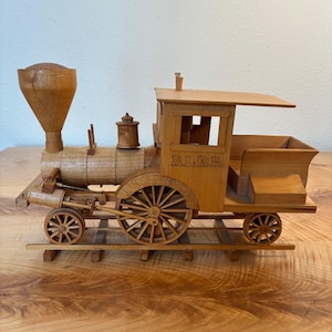 Puede incluir: Una locomotora de tren de madera con una gran chimenea en forma de embudo, una caldera cilíndrica y una cabina con el texto RLRR y PIONEER grabado en el lateral. El tren está montado sobre vías de madera con ruedas de radios.