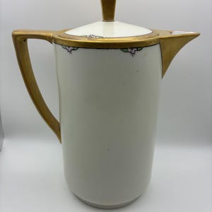 May include: A white porcelain coffee pot featuring a gold-painted handle, spout, and lid finial. The rim of the lid and the upper body are decorated with a delicate pattern of small pink flowers and green leaves, accented by thin black lines.