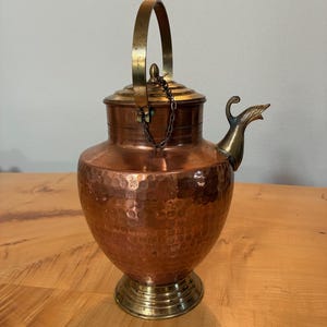 May include: A hammered copper vessel with a brass base, lid, and handle. The lid is secured to the handle with a small metal chain. The vessel features a curved brass spout and a wide, rounded body with a textured, dimpled surface finish.