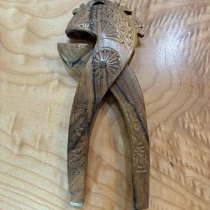 May include: A brown wooden nutcracker featuring intricate hand-carved geometric patterns, including sunburst designs on the circular pivot point and cross-hatch detailing along the two elongated handles and the curved upper jaw mechanism.