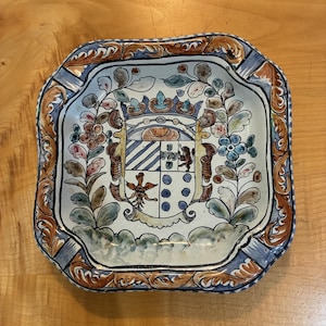 May include: A square ceramic dish with rounded corners, featuring a hand-painted central coat of arms topped with a crown. The design includes floral motifs in blue, green, and brown, with a decorative border of scrolling brown and blue patterns.