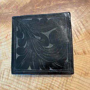 May include: A square black stone tile featuring an intricate, hand-etched floral pattern in a lighter gray tone. The design consists of sweeping, curved leaf-like shapes that fill the surface, framed by a thin, straight border line.