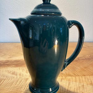 Op de afbeelding: Een donkergroenblauwe keramische koffiepot met een glanzende afwerking, voorzien van een bijpassend deksel met knop, een gebogen tuit en een stevig zijhandvat. De pot heeft een taps toelopend lichaam dat naar boven toe breder wordt op een ronde voet.
