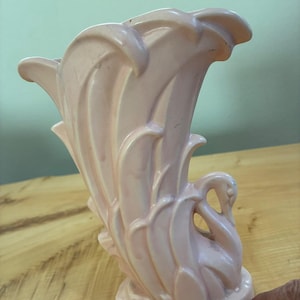 Puede incluir: Un jarrón de cerámica de color rosa pálido con forma de cisne, con plumas de ala estilizadas y superpuestas que forman el cuerpo y la abertura. La cabeza del cisne está situada en el lado derecho y hay una muesca visible en la base inferior.