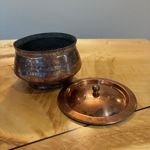 Peut inclure: Un pot en cuivre patiné avec un corps arrondi et un intérieur sombre, accompagné d'un couvercle circulaire en cuivre assorti doté d'un petit bouton en laiton. L'extérieur du pot présente des signes d'oxydation et une texture horizontale.