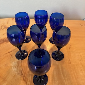 Puede incluir: Un conjunto de siete copas de cristal azul cobalto con cuencos redondeados, tallos delgados y bases circulares. Cada copa presenta un color azul profundo y uniforme con una superficie lisa y reflectante, dispuestas en un grupo compacto.