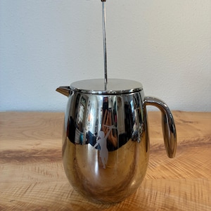 May include: A polished stainless steel French press coffee maker with a rounded body, a curved handle, and a vertical plunger mechanism. The exterior surface features a reflective finish with a small etched silhouette of a person holding an object.