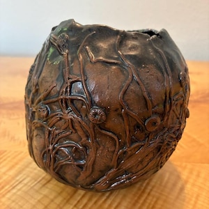 May include: A round, dark brown ceramic vase with an uneven, organic surface texture. The exterior features raised, root-like lines and small, circular, flower-shaped embellishments that wrap around the entire body of the vessel.