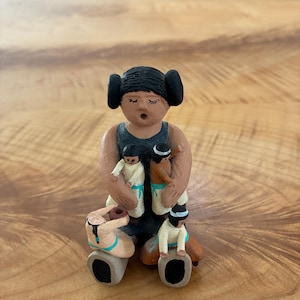 Peut inclure: Une figurine en argile représentant un adulte assis aux cheveux noirs en chignons, portant un vêtement gris foncé sans manches, tenant deux petits enfants, tandis que deux autres enfants sont assis à la base, l'un tenant un pot brun.