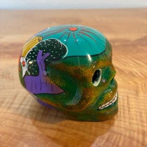 May include: A ceramic skull figurine featuring a glossy green and yellow mottled finish. The top and side are decorated with hand-painted designs including a teal circular pattern, a purple tree-like shape, and yellow and white floral accents.