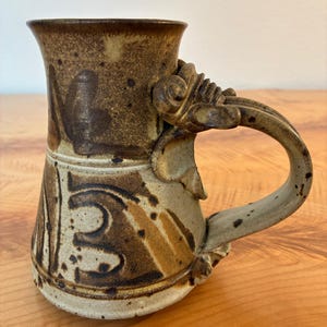 Może przedstawiać: Wysoki, cylindryczny kubek ceramiczny z nakrapianym beżowo-brązowym szkliwem. Kubek posiada ciemnobrązowe, abstrakcyjne wzory malowane pędzlem oraz wydatne, ozdobne ucho z rzeźbionym, zakręconym elementem dekoracyjnym w górnym punkcie mocowania.