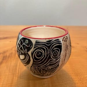 May include: A small ceramic cup with a cream-colored base, featuring a hand-carved black graphic pattern of swirling lines and abstract shapes. The rim of the cup is accented with a thin, vibrant red painted line, contrasting with the dark design.