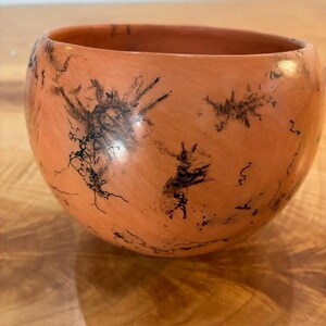 Może przedstawiać: Okrągła miska ceramiczna w kolorze terakoty o gładkim, matowym wykończeniu. Zewnętrzna powierzchnia jest ozdobiona misternymi, ciemnoczarnymi, rozgałęzionymi wzorami przypominającymi paprocie, które oplatają zakrzywiony korpus naczynia.