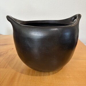 May include: A matte black ceramic bowl with a rounded, bulbous body and two integrated handles extending from the upper rim. The vessel features a smooth, dark finish and a wide, open top, showcasing a simple, functional design with no additional patterns.