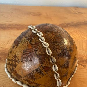 May include: A rounded, tan-coloured gourd vessel featuring a smooth, polished surface decorated with intricate, hand-etched geometric patterns in dark brown. A single strand of white cowrie shells is draped across the top and around the base.