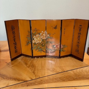 May include: A six-panel folding screen with a glossy amber finish, featuring painted floral motifs of white and pink peonies, green leaves, and a small butterfly. The outer panels display black Chinese calligraphy characters on the smooth surface.