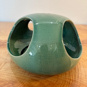 May include: A sage green ceramic bowl with a rounded, organic shape and a distinct crackle glaze finish. The bowl features three large, arched cutouts along its sides, revealing a smooth, darker green interior surface and a hollow center.
