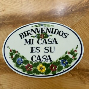 May include: An oval ceramic wall plaque with a white background and a dark blue border. The text "BIENVENIDOS MI CASA ES SU CASA" is printed in black. The plaque is decorated with hand-painted green leaves and colourful flowers in red, yellow, and blue.