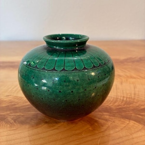 May include: A small, round, green ceramic vase with a glossy finish. The upper section features a decorative band of dark, petal-like shapes, while the lower body is adorned with a repeating pattern of small, dark, evenly spaced dots.