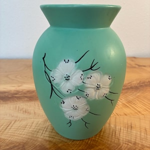 May include: A mint green ceramic vase with a wide, flared rim and a rounded body. The front of the vase features a hand-painted design of three white flowers with black centers, connected by thin, black, hand-painted branches and stems.