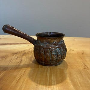 May include: A copper coffee pot with a rounded, textured body featuring vertical indentations and a dark, weathered patina. It has a long, dark brown wooden handle attached to the side, which is secured with visible metal rivets along its length.