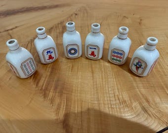 Puede incluir: Seis botellas pequeñas de cerámica blanca con forma de fichas de mahjong, cada una con un gráfico colorido único en el frente. Las botellas tienen un cuello estrecho y un marco rectangular con borde dorado alrededor del símbolo central.
