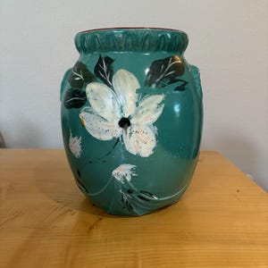 Op de afbeelding: Een blauwgroene keramieken vaas met een geschulpte rand en een handgeschilderd wit bloemmotief met donkergroene bladeren. De vaas heeft een glanzende afwerking en een ronde lichaamsvorm met kleine handvatten aan de zijkanten.