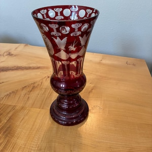 May include: A deep red glass vase featuring intricate white etched designs, including circular patterns, stylized birds, and architectural motifs. The vase has a flared rim, a bulbous midsection, and a circular pedestal base with detailed engravings.