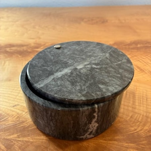 Puede incluir: Un recipiente redondo de mármol gris oscuro con una tapa circular a juego. La piedra presenta vetas blancas naturales en toda la superficie. Un pequeño acento metálico circular está incrustado cerca del borde de la tapa, que está desplazada.